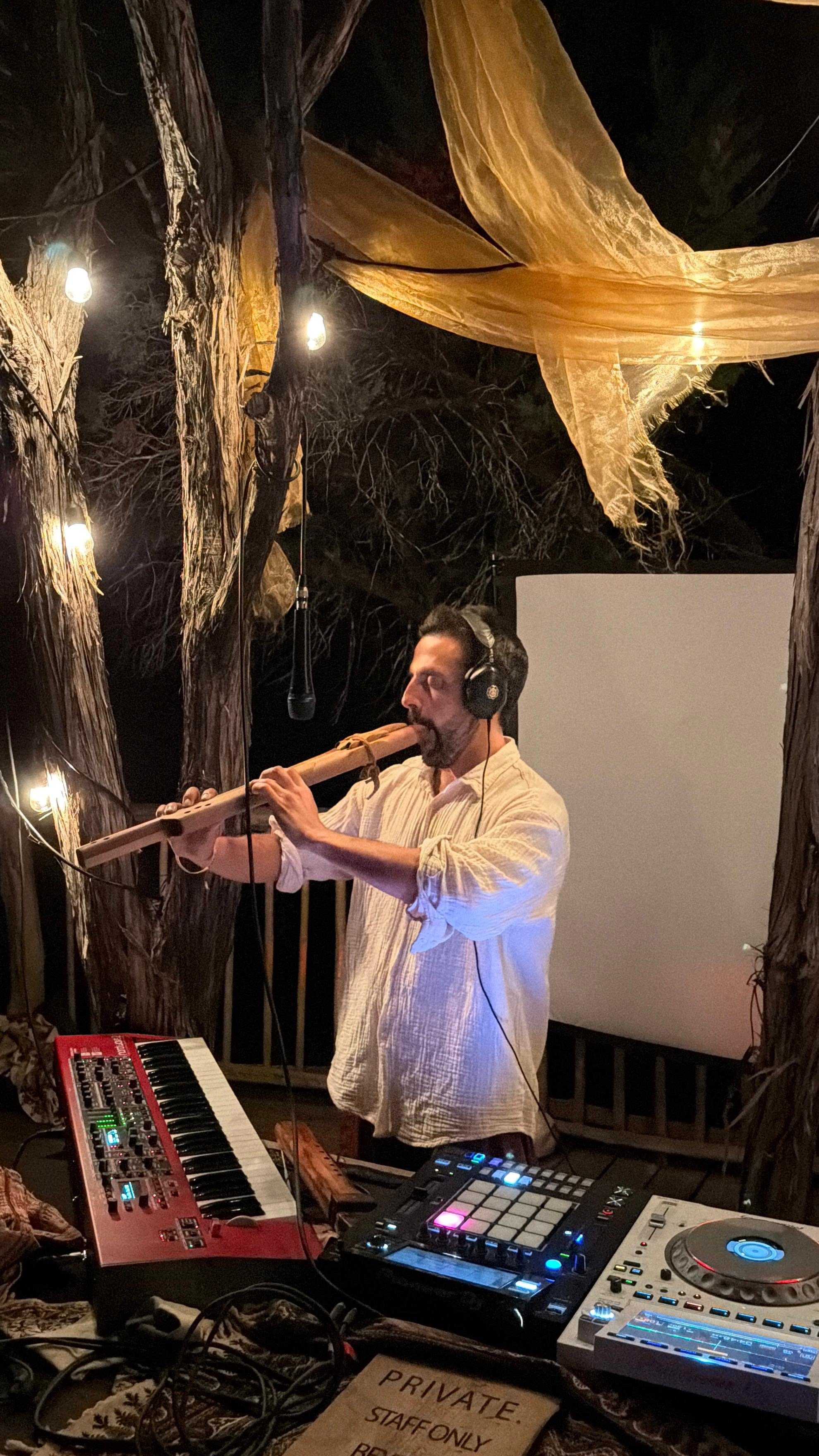 The artist Kora playing wooden flute over synths under gold drapes and string lights
